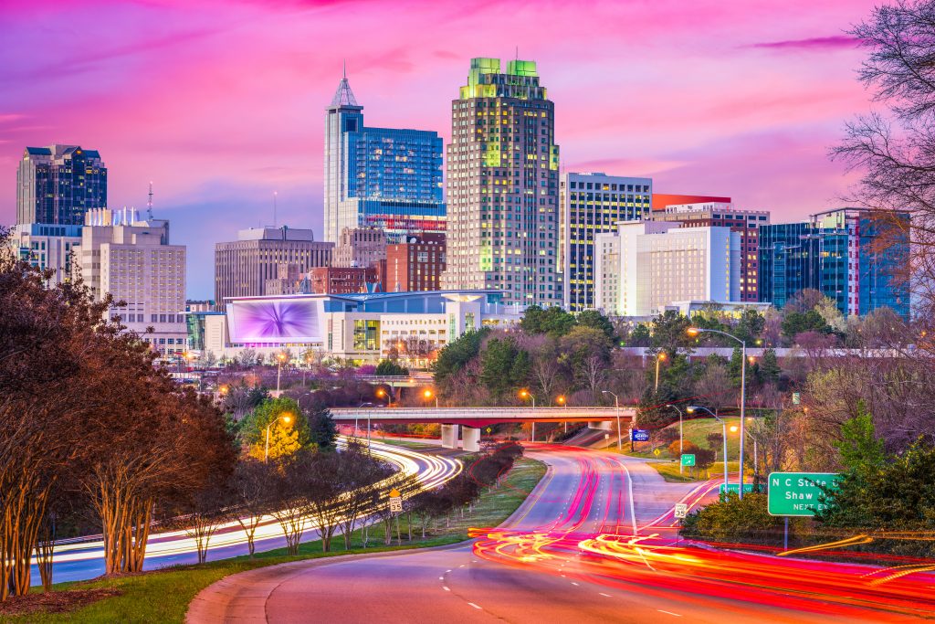 City skyline at dusk with vibrant pink and blue sky. Tall buildings are illuminated, and light trails from vehicles create dynamic lines on a highway in the foreground. Green and brown foliage borders the road. A sign reads N. St. Marys St.