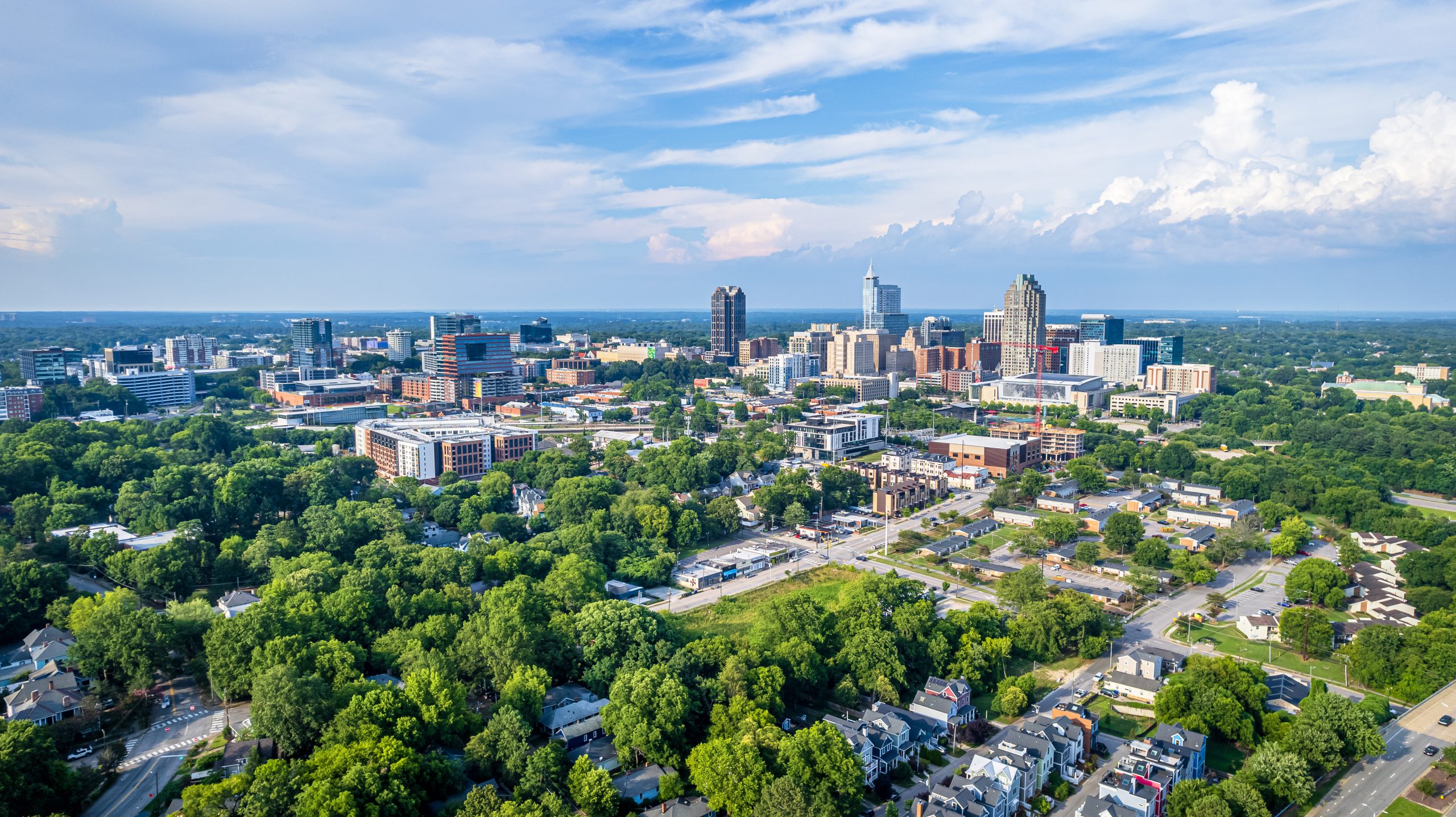 Aerial view of a cityscape showcasing a lush green landscape with numerous trees. The city skyline features a mix of modern and traditional buildings under a blue sky with scattered clouds. Residential and urban areas are visible.