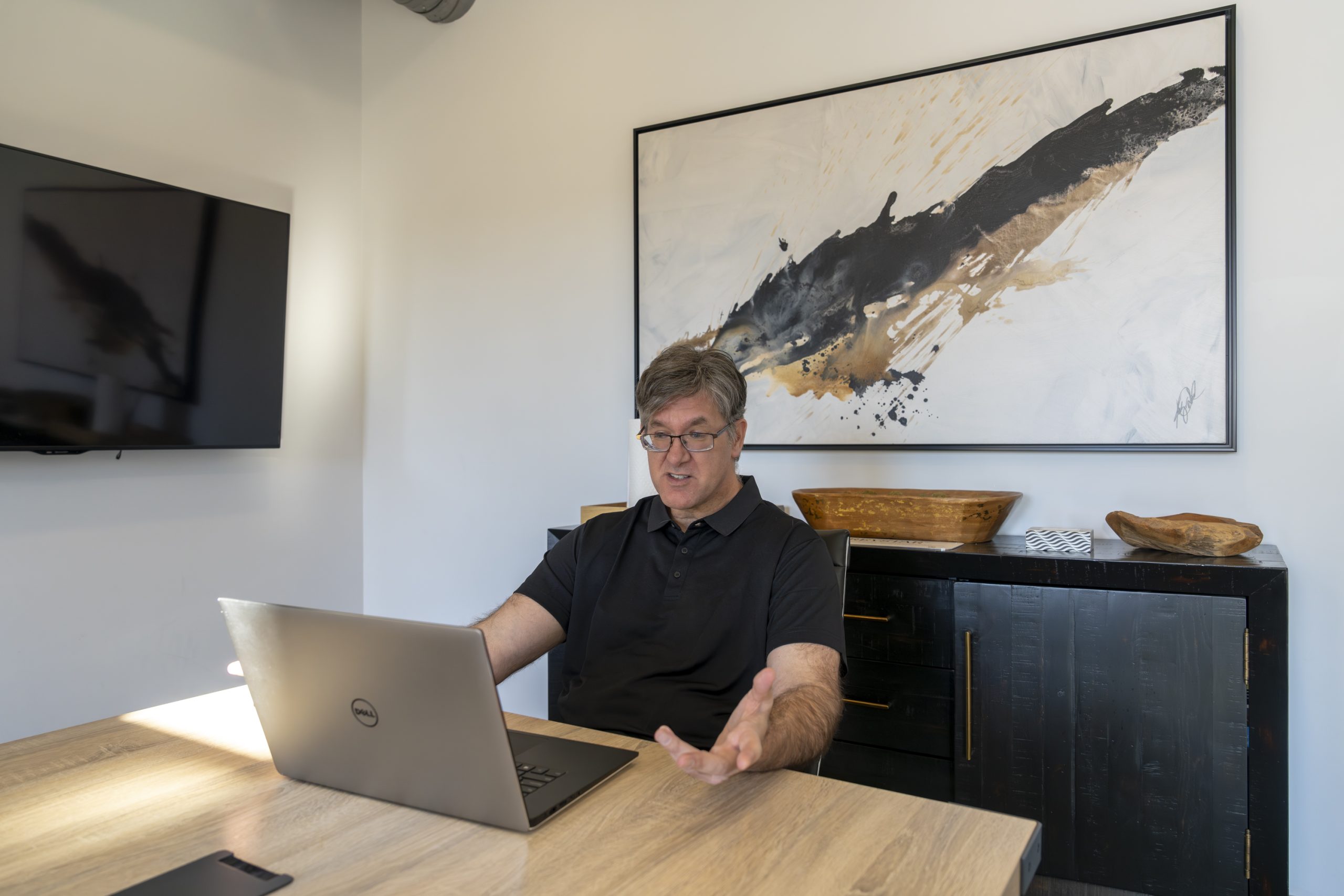 A person is sitting at a desk using a laptop in a modern office. They appear to be talking or presenting, with one hand gesturing. Behind them is a large abstract painting and a wooden cabinet with decorative items.