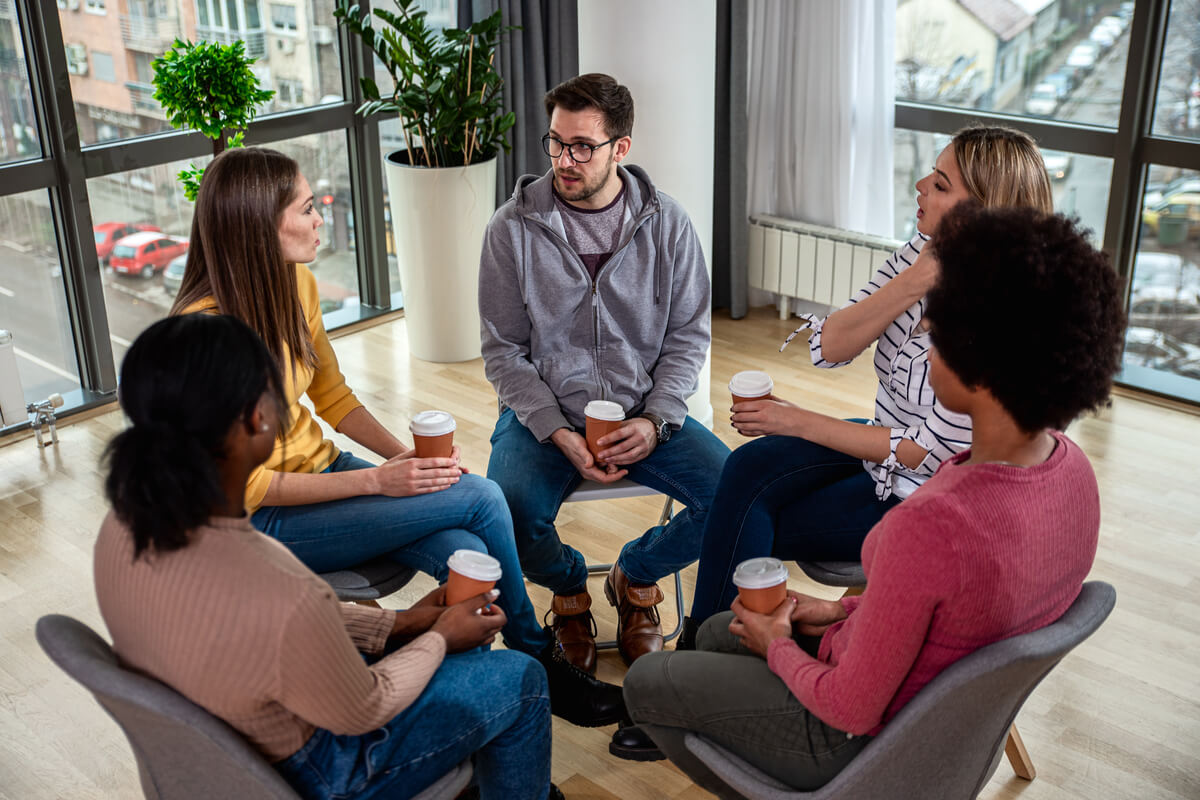 Five people sitting in a circle in a bright room, holding coffee cups and engaging in conversation. The group consists of two men and three women, all casually dressed. Large windows reveal an urban street and buildings outside.