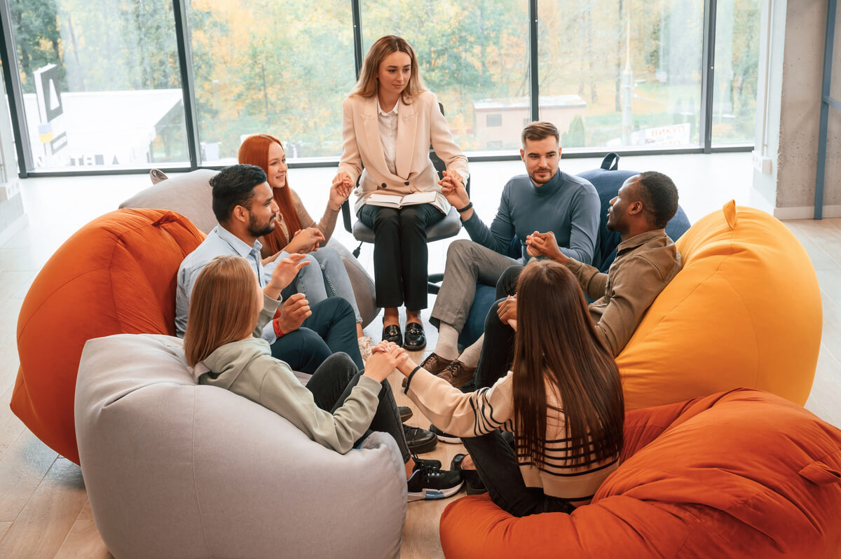 A group of seven people sit in a circle on large bean bags, holding hands. They appear to be engaged in a group activity indoors, with large windows showing trees in the background. The atmosphere seems relaxed and collaborative.