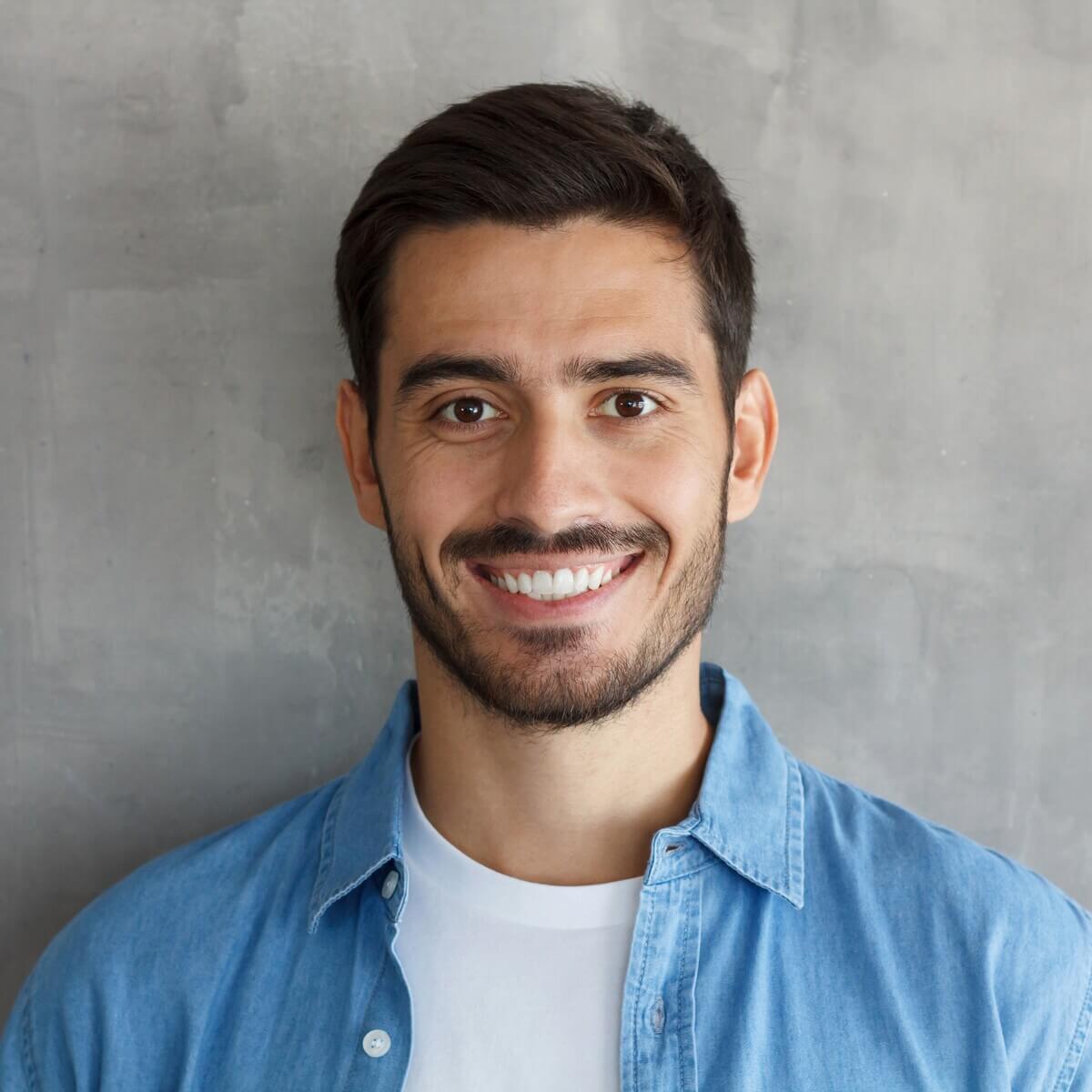 Smiling man with short dark hair and a beard, wearing a blue denim shirt over a white t-shirt, standing against a textured gray background.