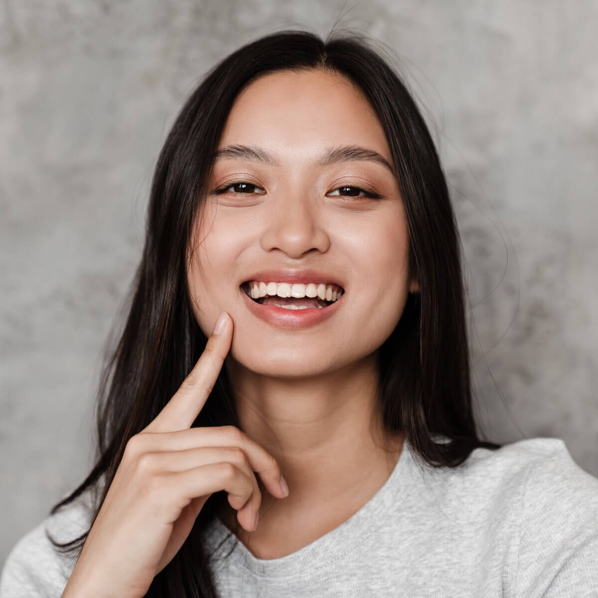 A person with long, dark hair is smiling, looking directly at the camera. They are wearing a light gray top and have one hand gently touching their face. The background is a plain, textured gray.