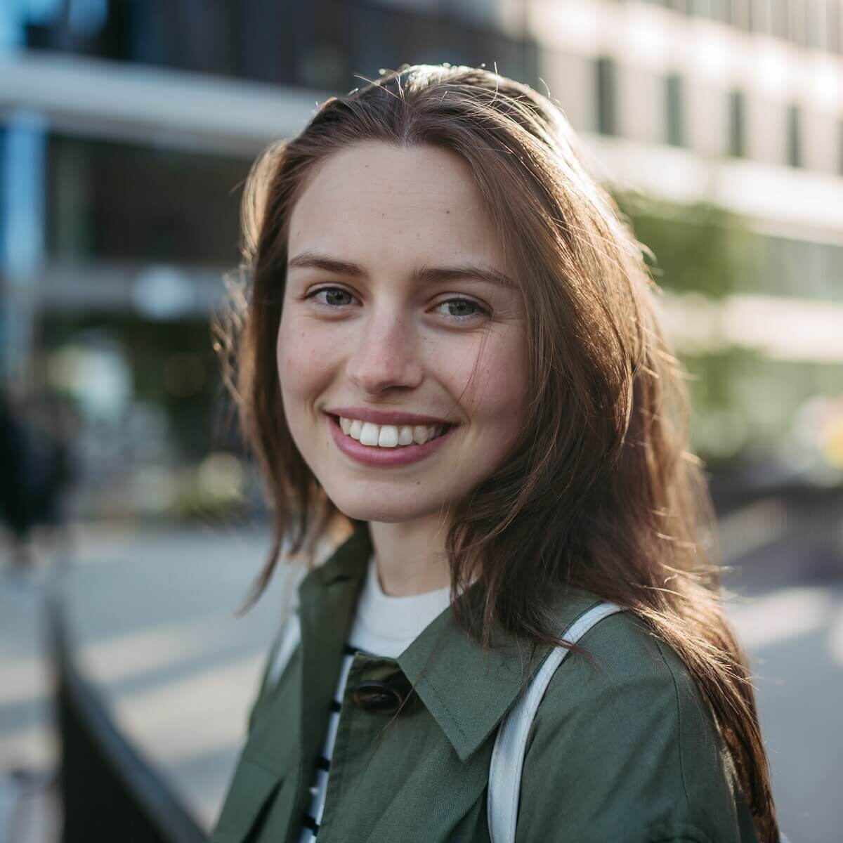 A person with long brown hair smiles warmly at the camera. They are outdoors in an urban setting, wearing a green jacket. The background is softly blurred, showing buildings and greenery in the sunlight.