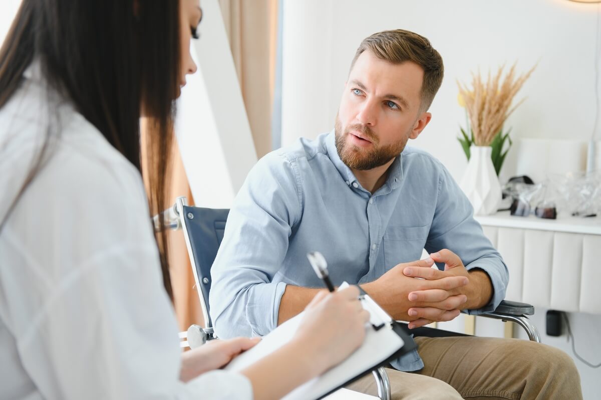 A man with a beard is sitting and talking to a woman with long dark hair holding a clipboard. They appear engaged in a conversation in a bright room with a white table and decorative vase in the background.