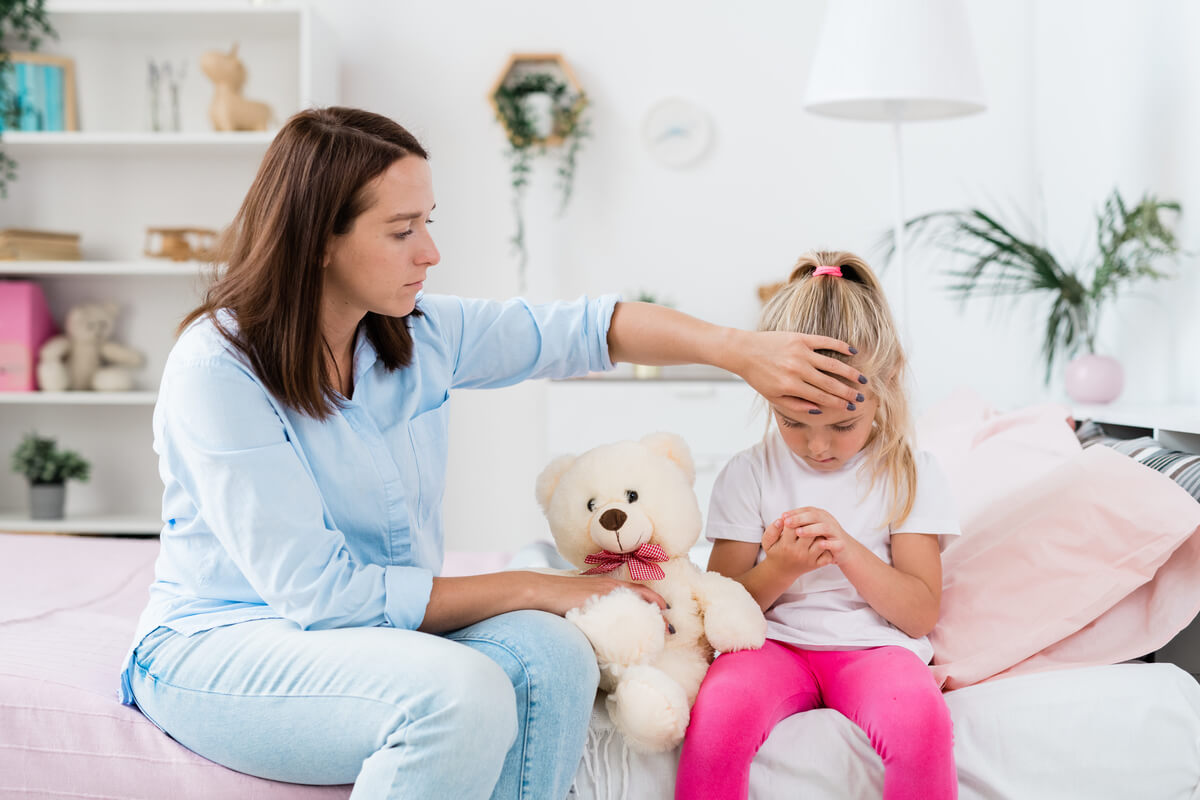 A woman in a blue shirt gently touches the forehead of a young girl sitting on a bed with a teddy bear. The girl, wearing a white shirt and pink pants, looks down at her fingers. The room has a cozy and bright decor with plants and shelves.